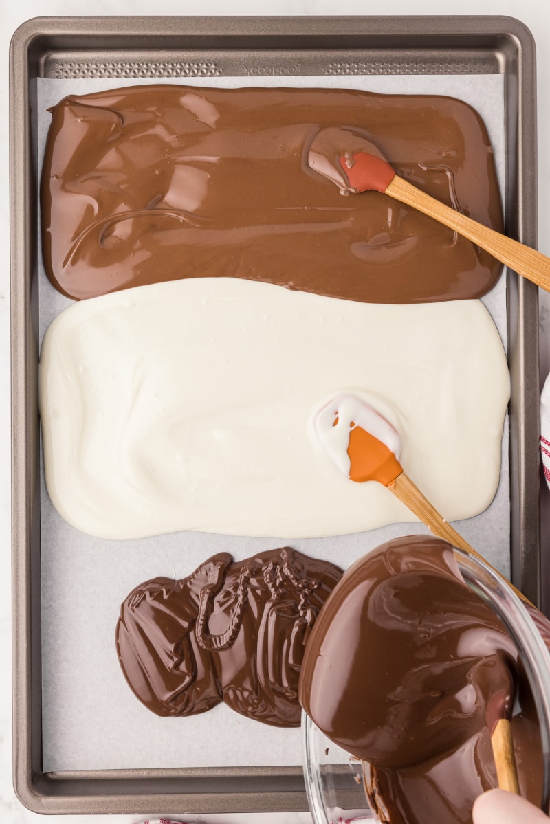 A metal cookie sheet covered in parchment paper with dark and white melted chocolate spread out with a spatula