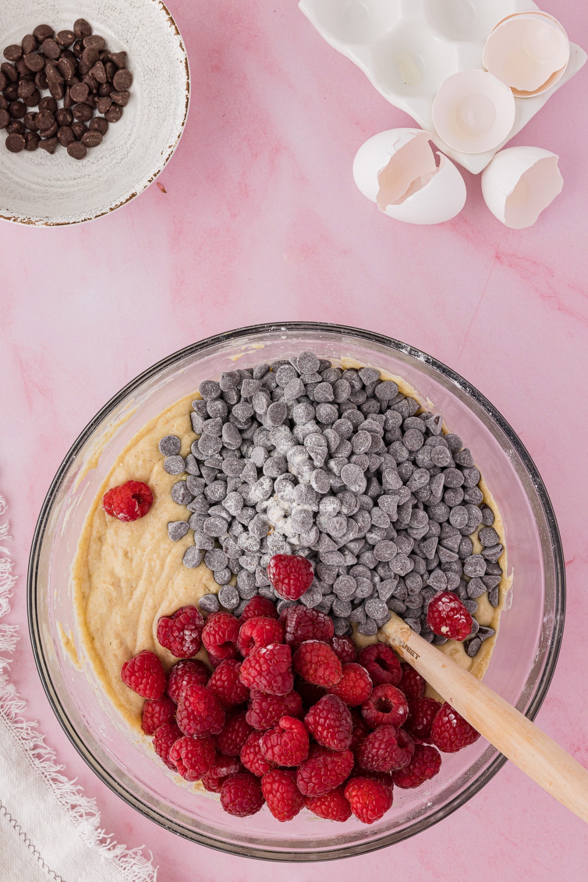 Muffin batter in a bowl with raspberries and chocolate chips on a pink table top