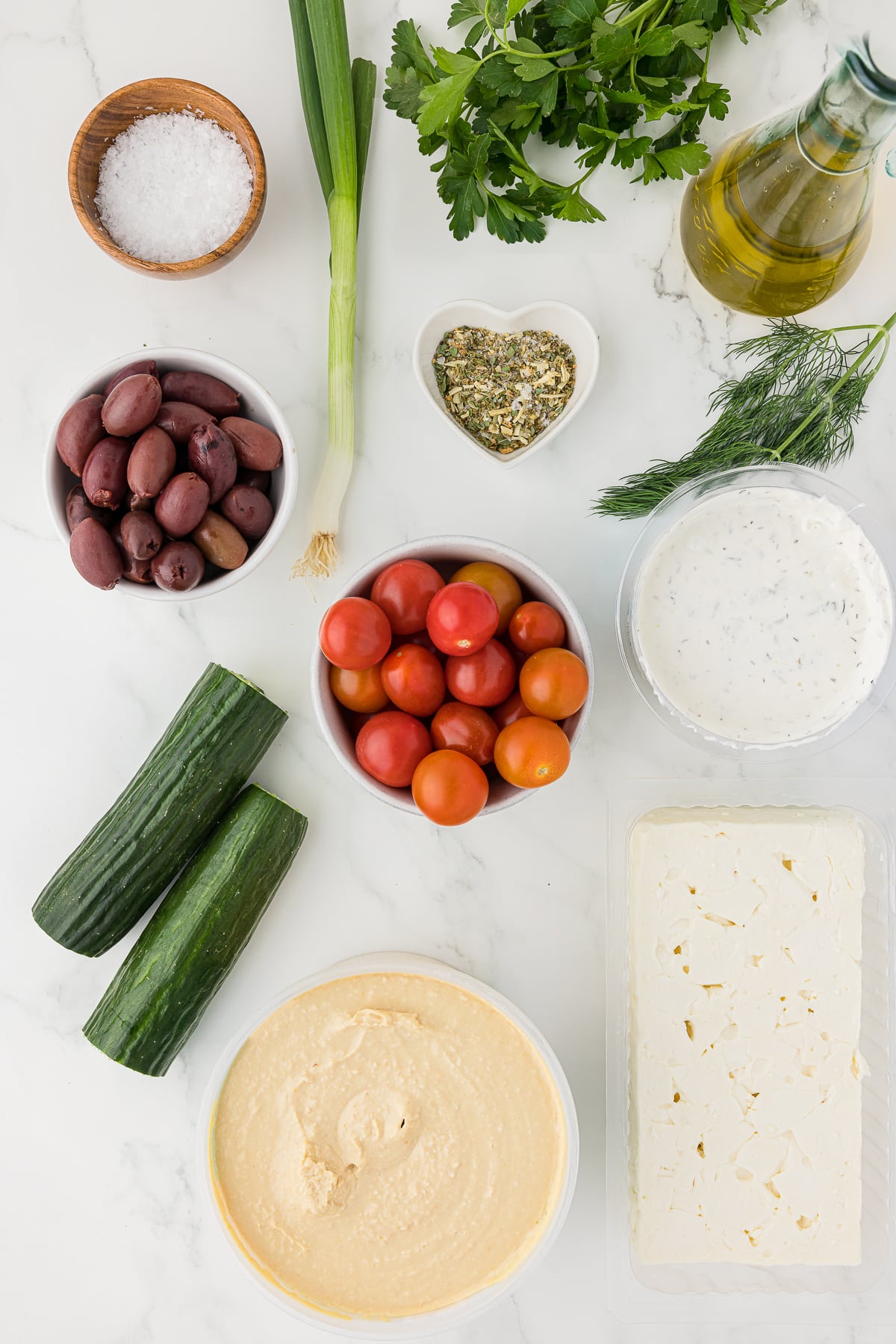Several ingredients for a greek party dip recipe all spread out in their containers on a white counter top