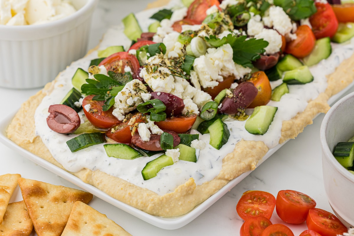 A party tray full of a greek appetizer dip topped with feta cheese with crackers for dipping