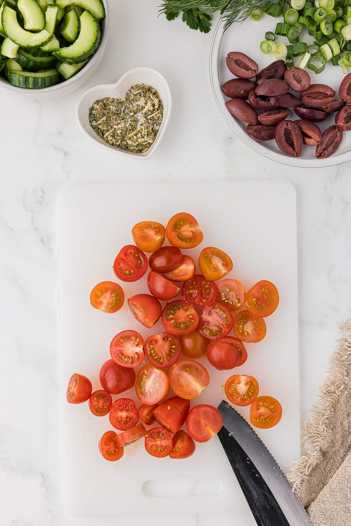Sliced cherry tomatoes on a cutting board surrounded by olives, onions, cucumbers and spices