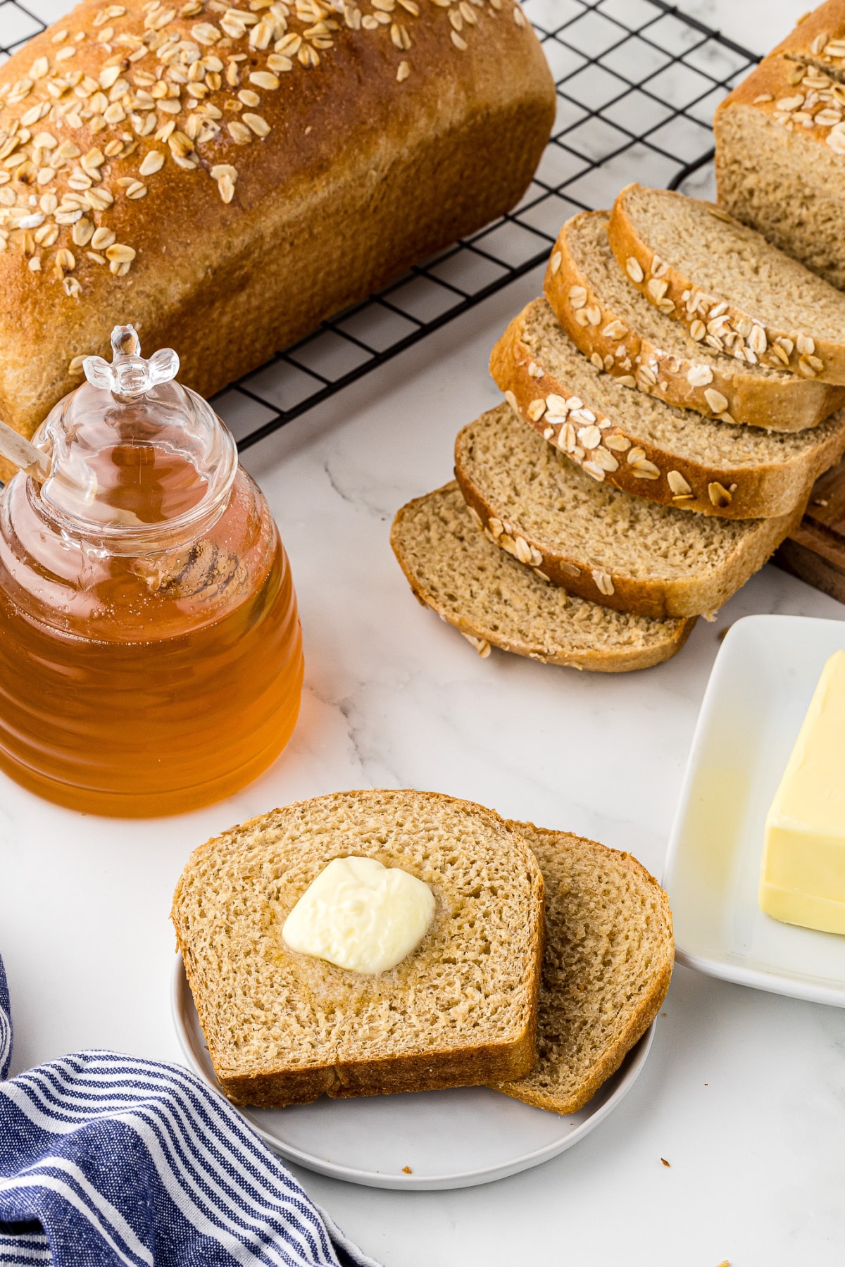 Two slices of bread on a plate with butter on top and a jar of honey and loaf of bread in the background