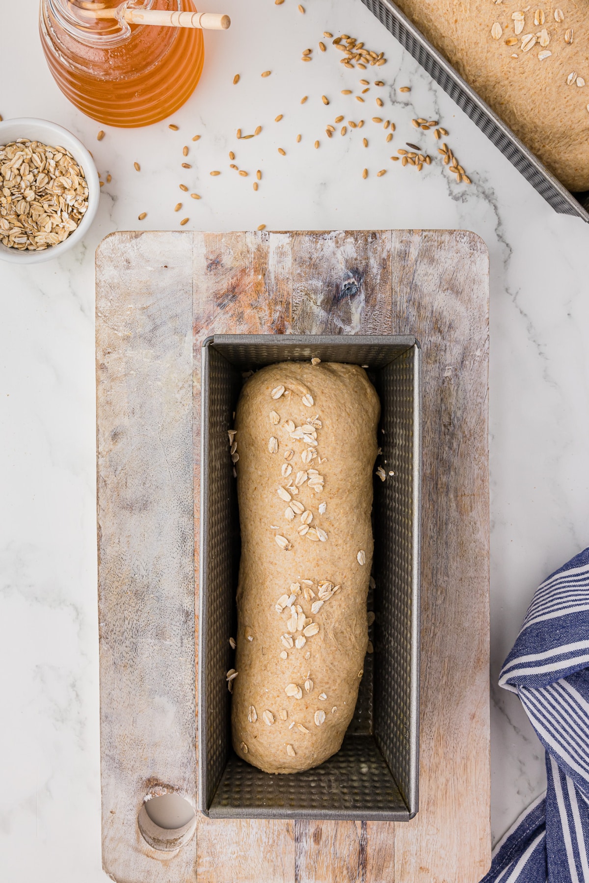 Bread dough shaped into a bread pan on a wooden cutting board with honey and oats laying on the counter