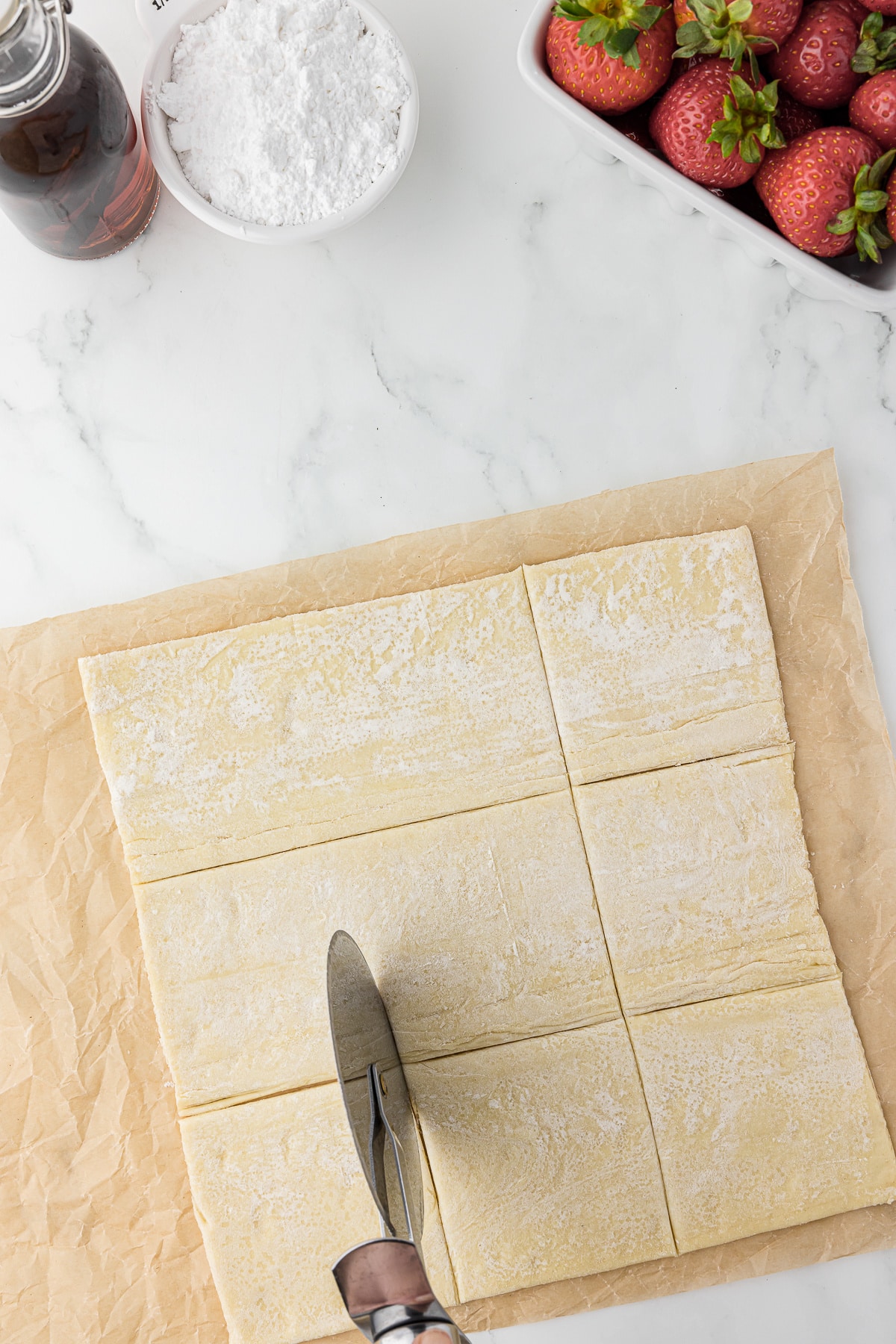 A pizza cutter cutting up squares in a sheet of puff pastry with strawberries and powdered sugar in the background