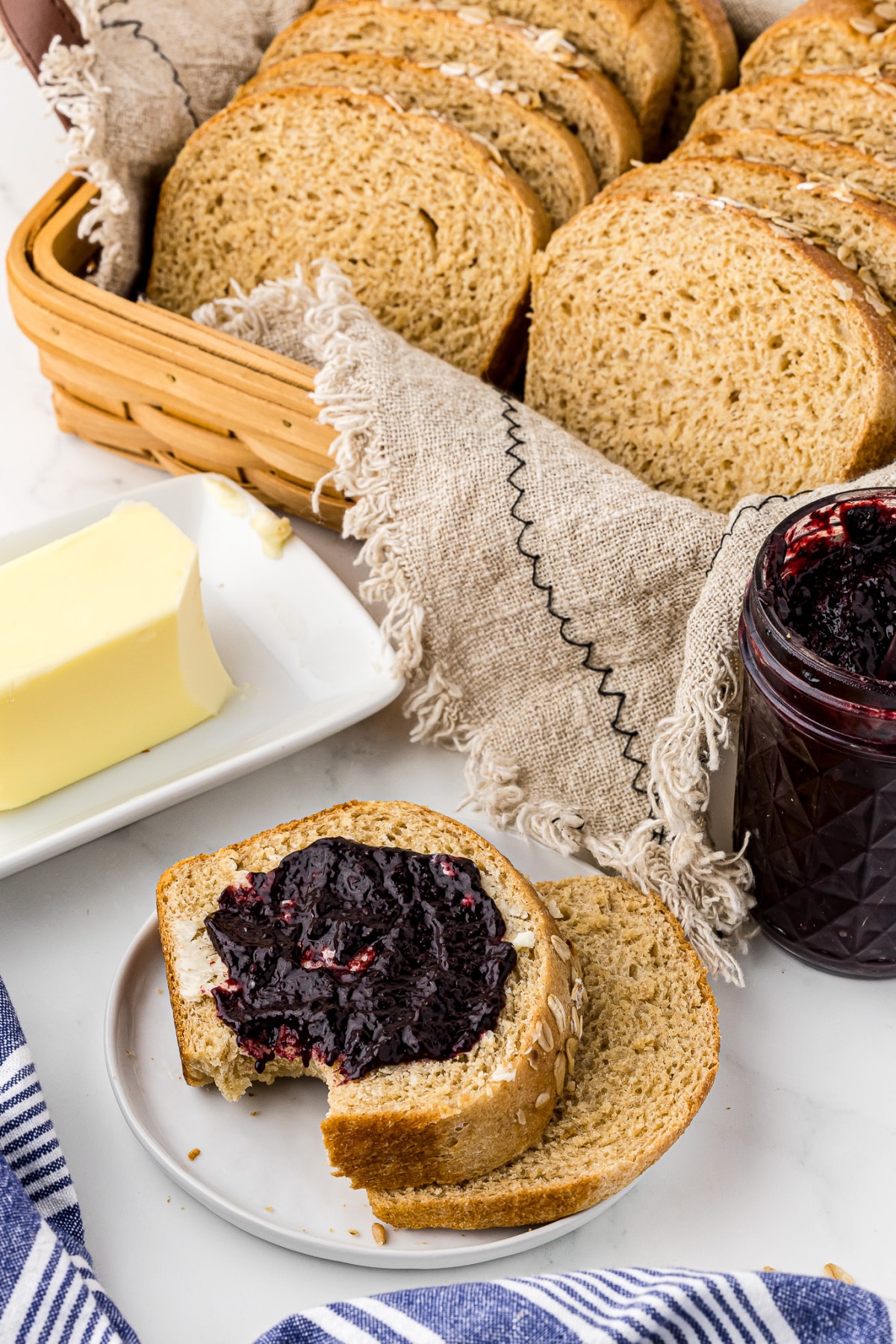 Slices of bread on a white dish with jam topping and a bread basket full of bread along with a jar of jam and a butter dish in the background