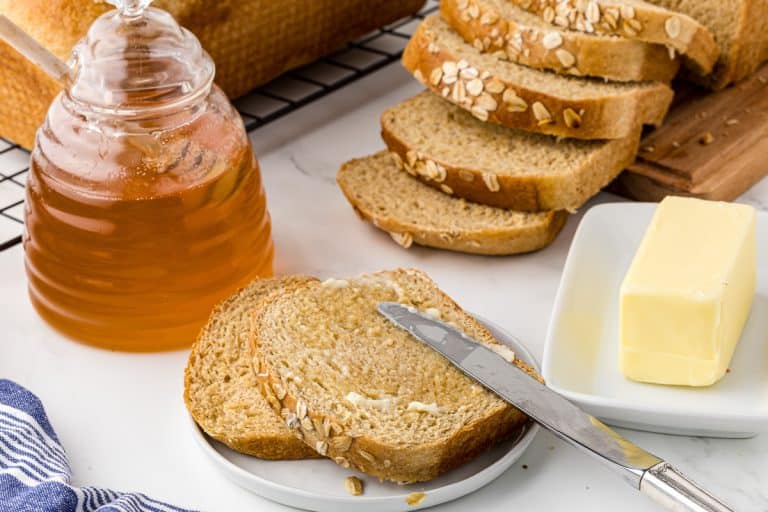 Sliced toast on a plate with a butter knife and dish of butter and a jar of honey and sliced loaf of bread in the background