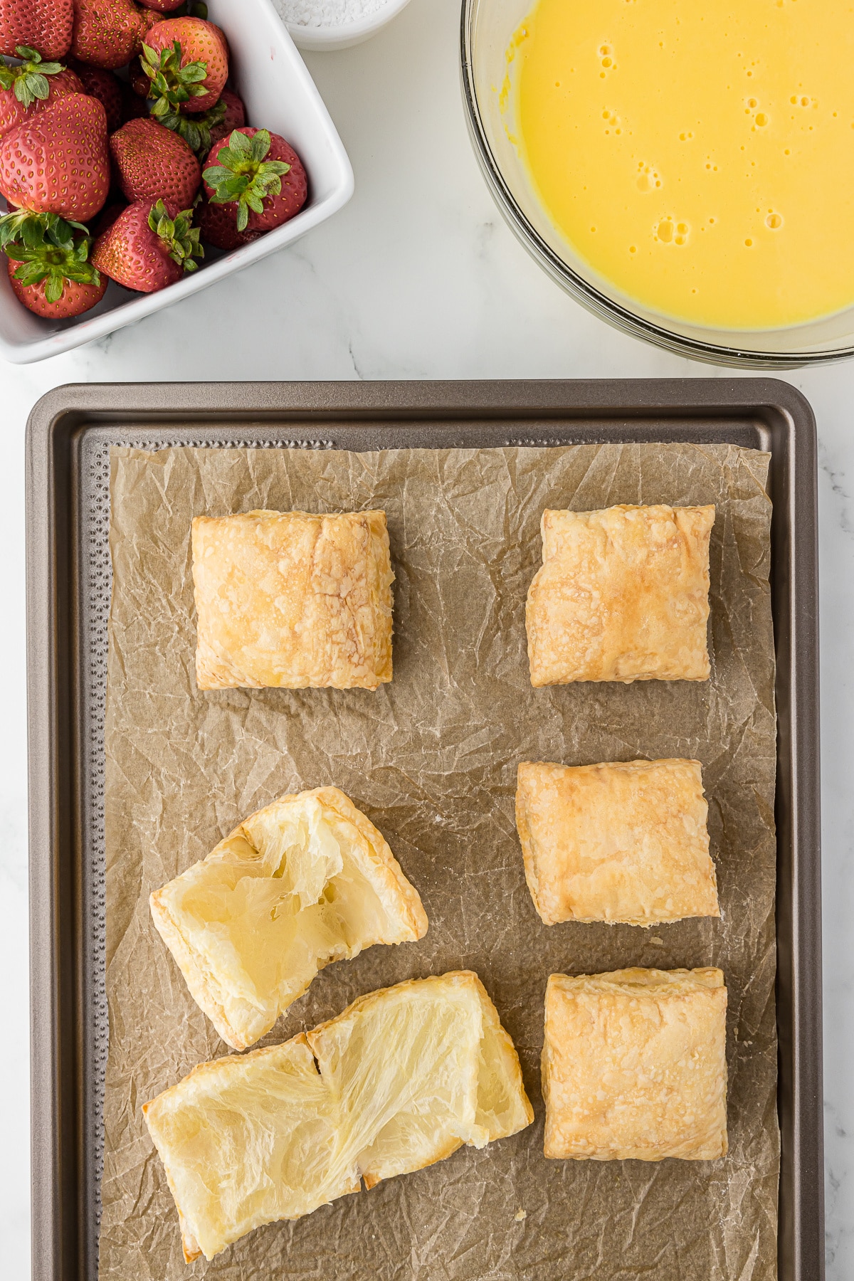 Puffed pastry squares on a baking sheet with parchment paper and a bowl of strawberries and custard in the background