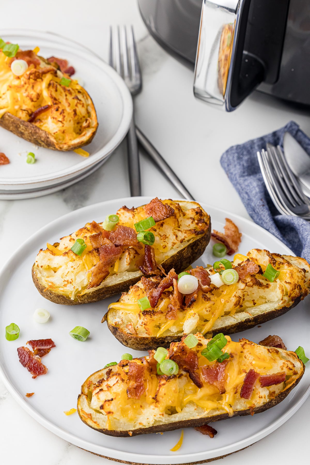 Three baked potatoes on a serving plate on a white countertop with an air fryer and forks and knives in the background