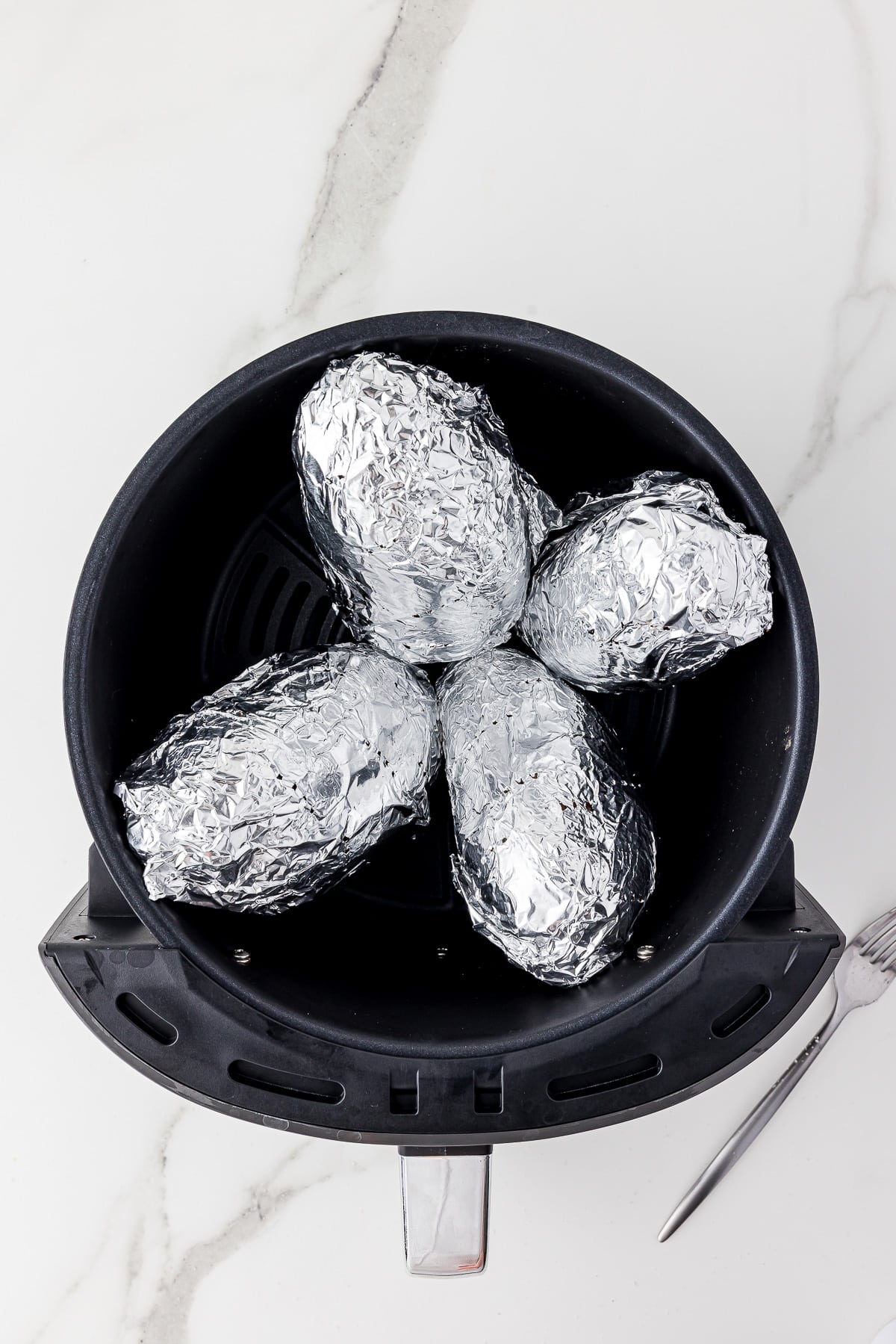 four foil covered russet potatoes in an air fryer basket sitting on a white marble counter