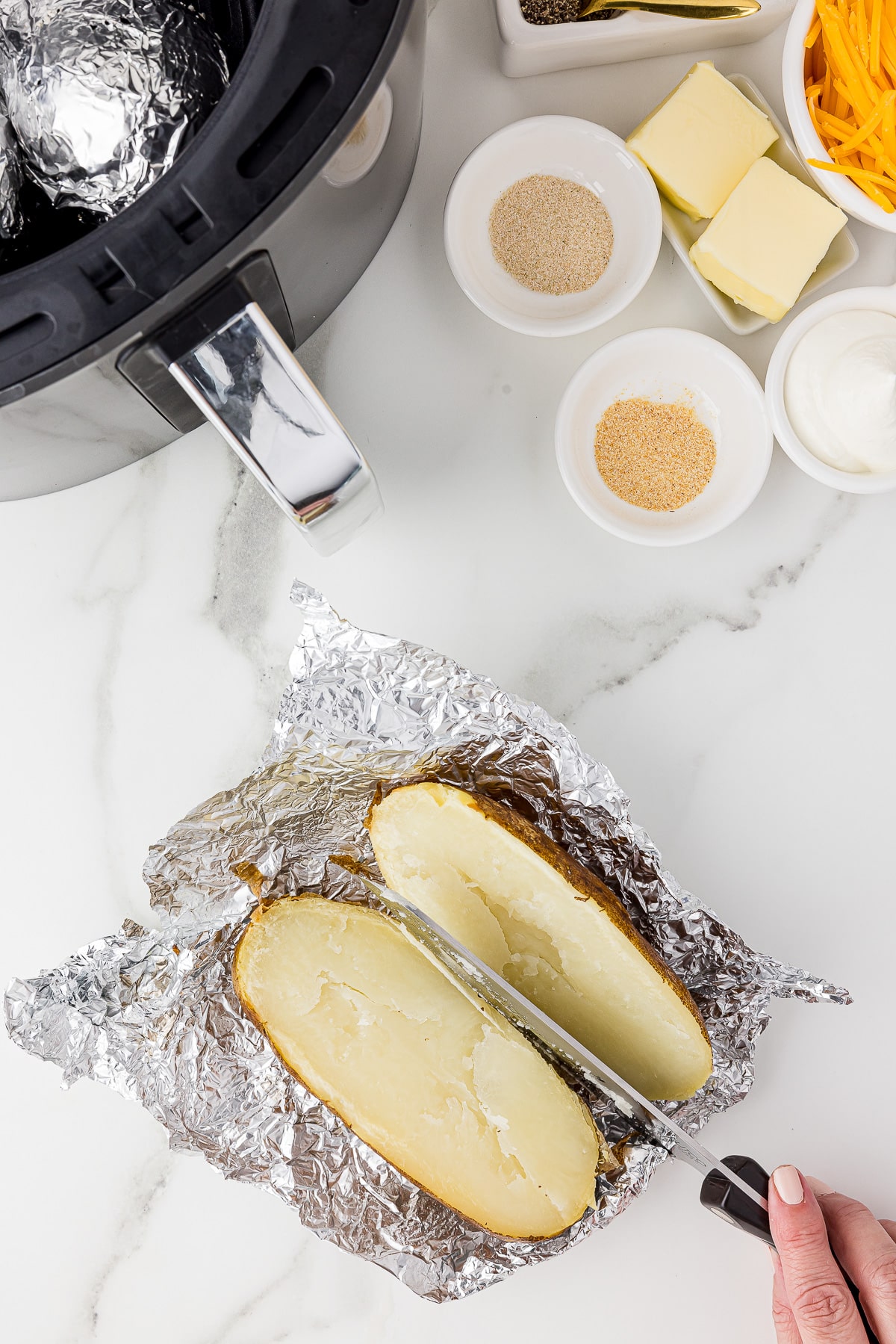 a baked potato being cut in half on aluminum foil on a white marble counter, pinch bowls of ingredients and an airfyer in the backgroun