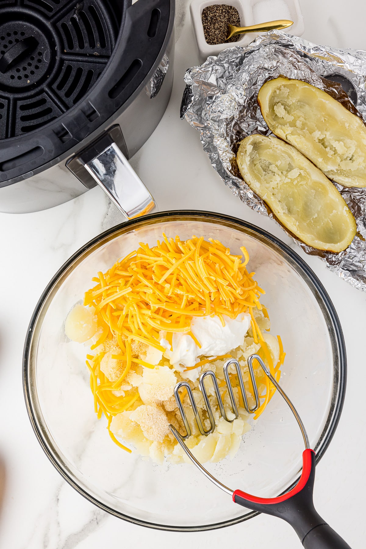 potato masher in a bowl with shredded cheese, sour cream and potato pulp on a white counter top alongside an air fryer and empty shells