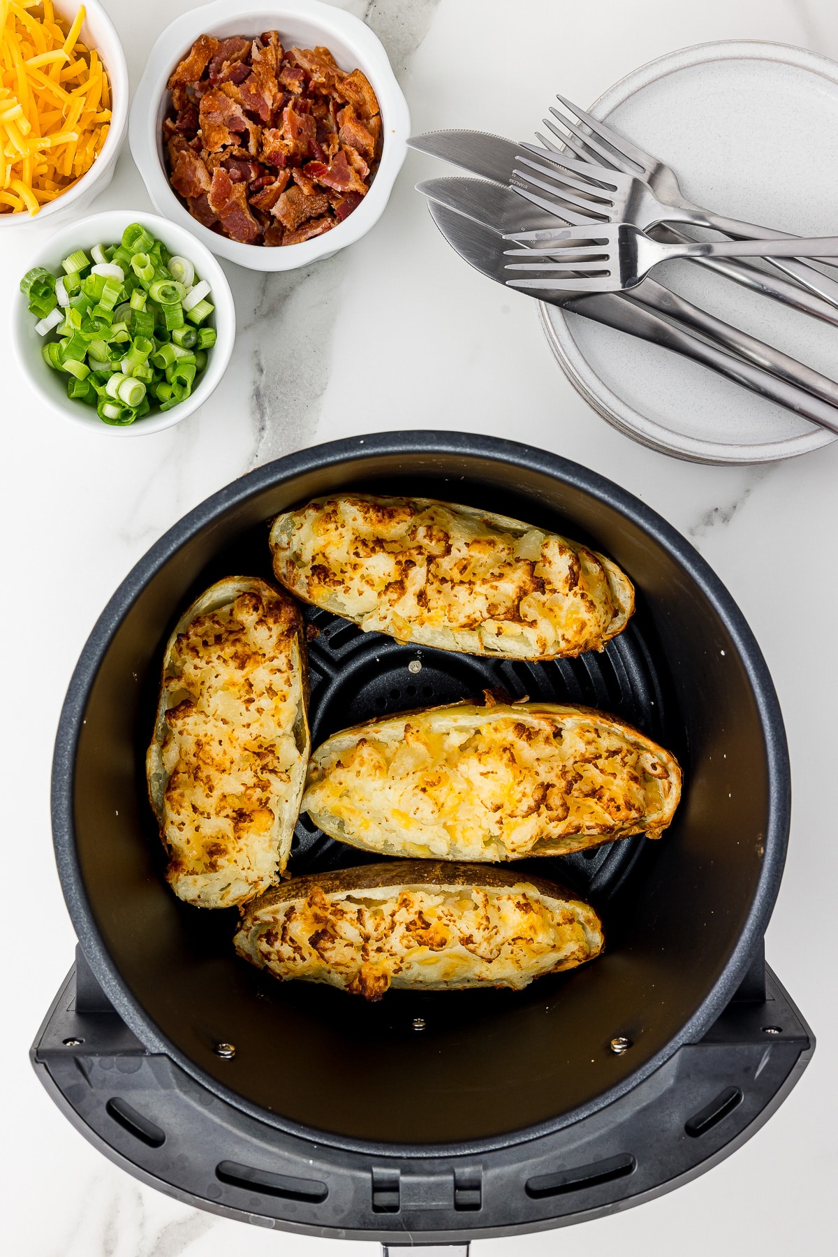 four twice baked potatoes in an air fryer bowl on a white counter with smallserving bowls of bacon, green onions, and shredded cheddar cheese