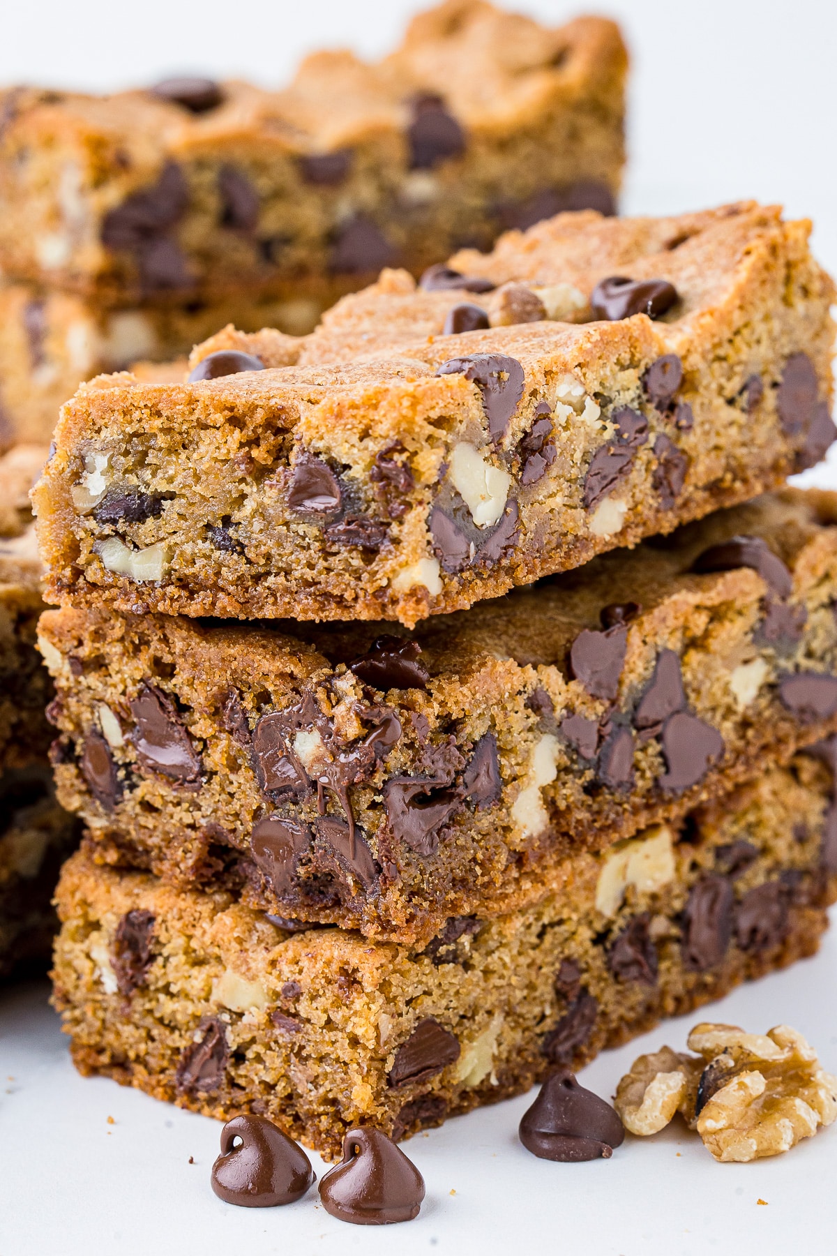 closeup of chocolate chip cookie bars with walnuts and chocolate chips in the foreground, all on a white marble countertop.