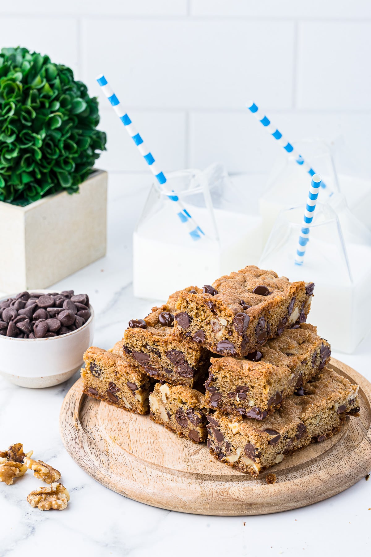 chocolate chip cookie bars on a wooden platter with milk in glass jars, a plant, and chocolate chips in the background, all on a white marble countertop