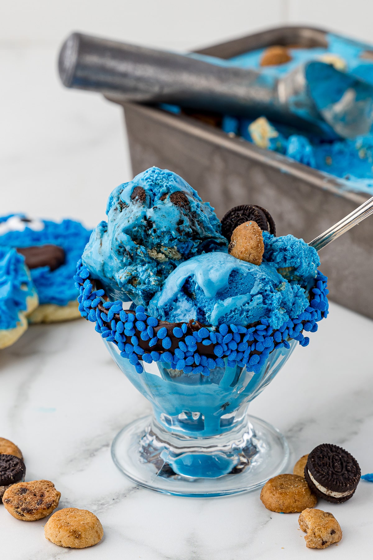 Blue cookie monster ice cream in a glass sunday with blue sprinkles on a white marble countertop with cookie monster sugar cookies and a loaf pan filled with blue ice cream in the background