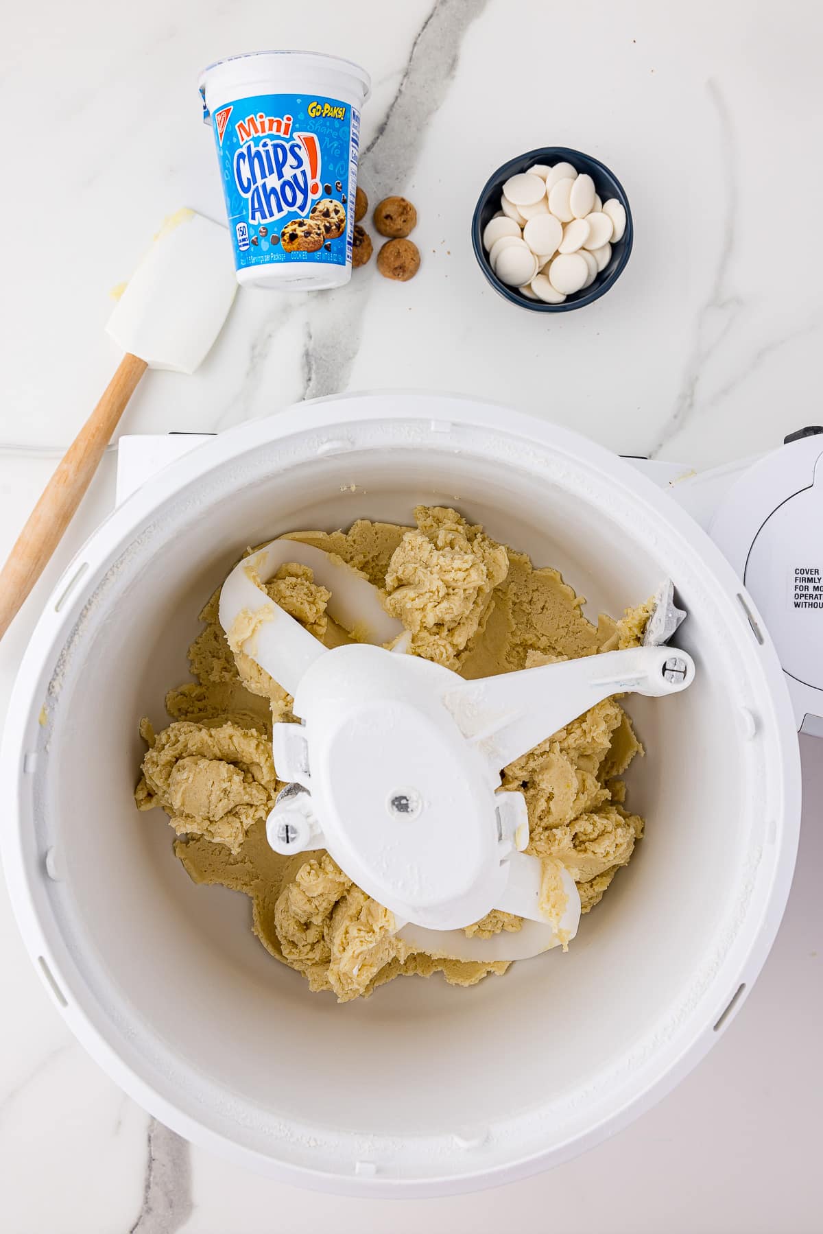 sugar cookie dough being mixed in a bosch mixer on a white marble countertop with mini chips ahoy cookies and white melting discs in small blue bowl and a white spatula