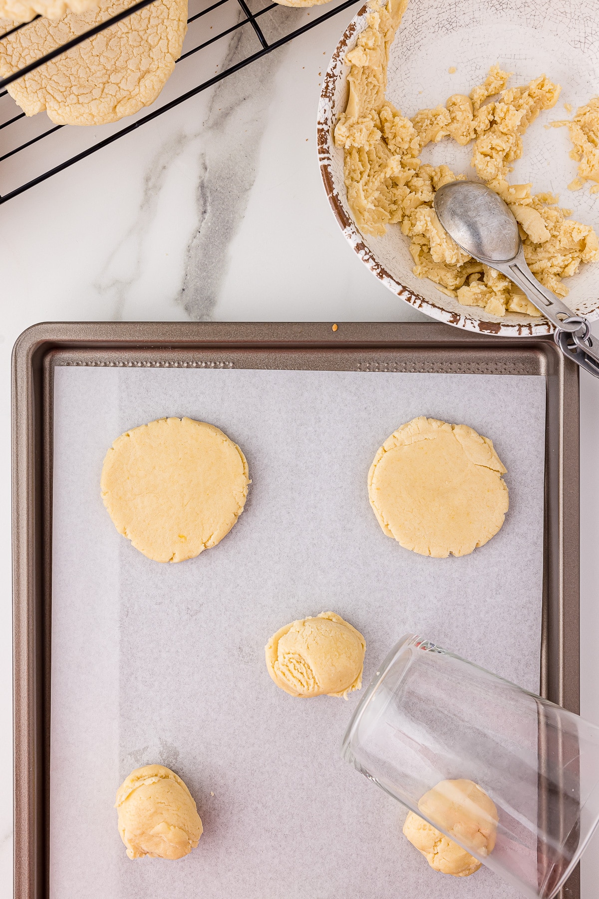 cookie dough being flattened with the bottom of a glass on a cookie sheet with a bowl of raw dough and sugar cookies cooling on a wire cooling rack