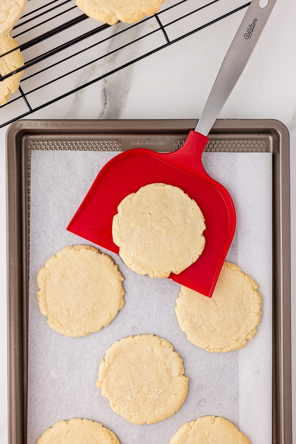 6 baked sugar cookies on a cookie sheet, one cookie being scooped up with a wilton red oversized spatula and sugar cookies cooling on a wire cooling rack