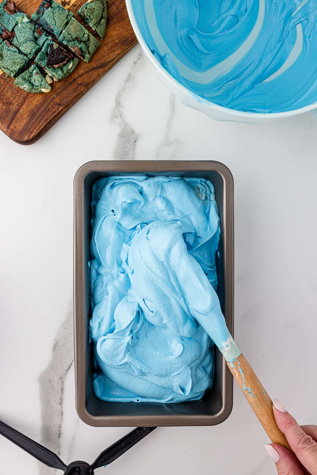 Blue ice cream being spread into a metal bread pan with a spatula