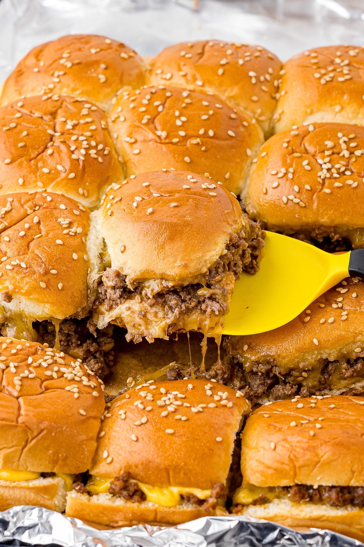 a tray of Hawaiian rolls cheese sliders, one being lifted out of the pan with a yellow spatula