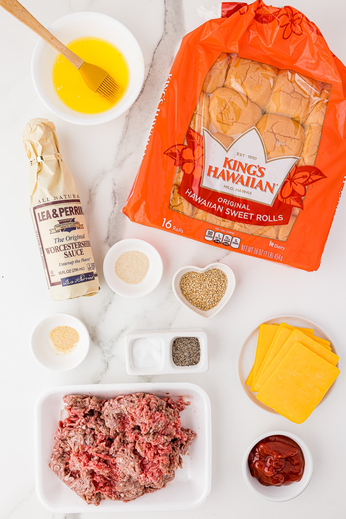 Ingredients on a white marble countertop, including King's Hawaiian sweet rolls, melted butter, ground meet, ketchup, and sliced cheddar cheese