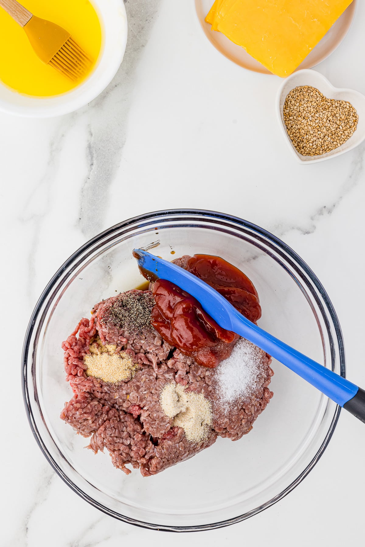 clear glass mixing bowl with raw ground meat, ketchup, spices and a blue spatula on a white marble countertop, with sliced cheese, sesame seeds and melted butter in the background