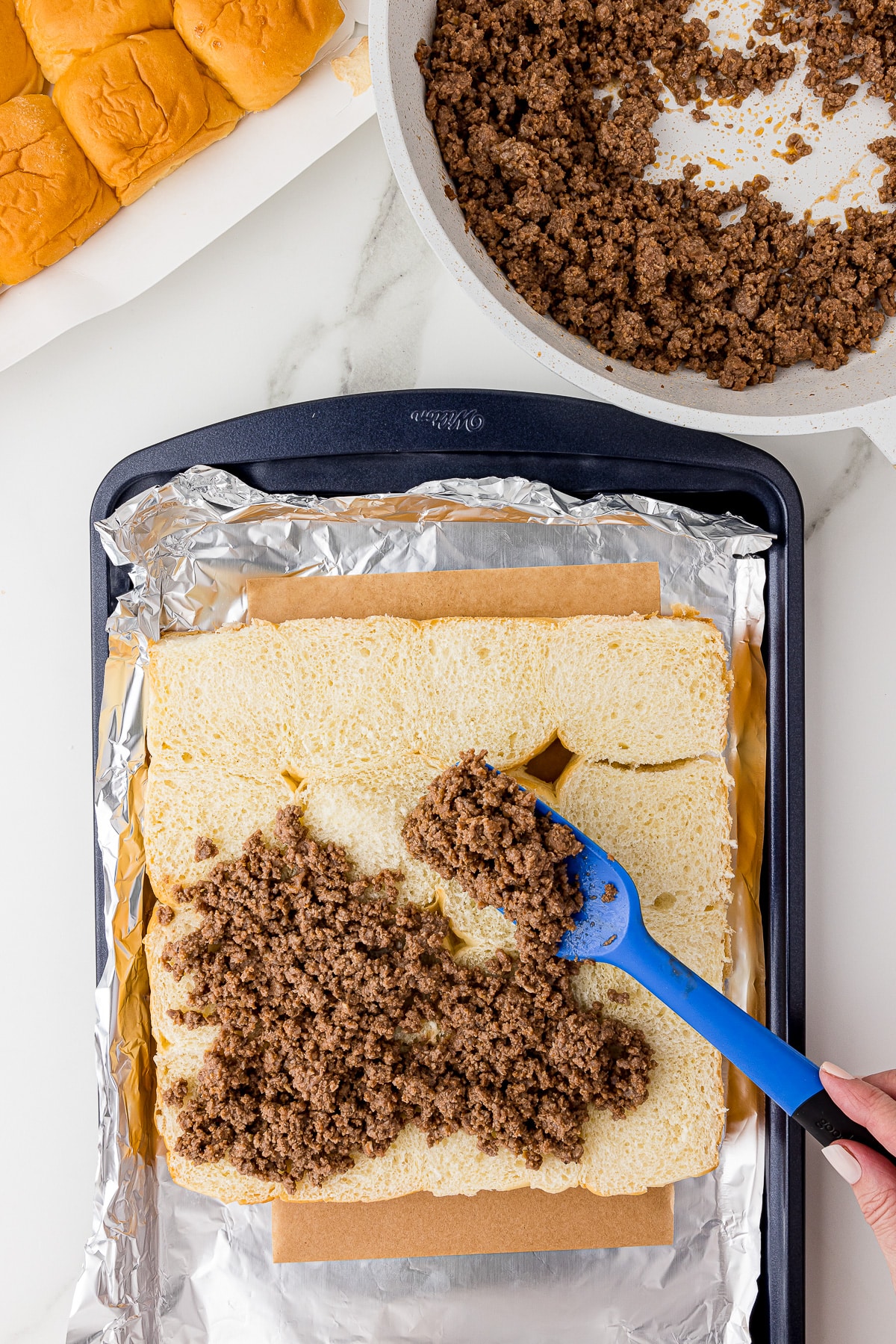 blue spatula spreading cooked ground meat onto Hawaiian rolls in a navy cookie sheet lined with foil, rolls and a frying pan with more cooked hamburger in the back