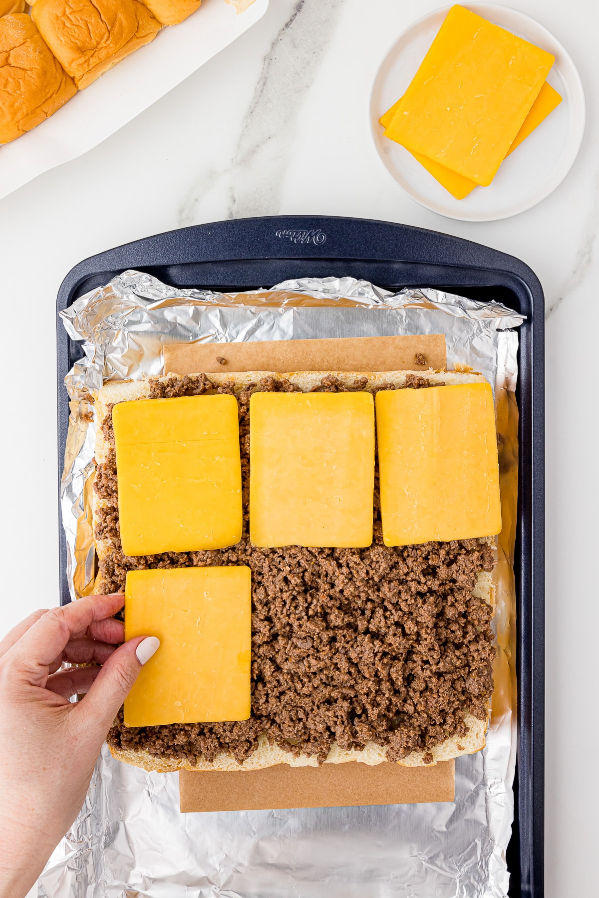 placing slices of cheddar cheese on cooked hamburger in a Wilton cookie sheet lined with foil on a white marble countertop