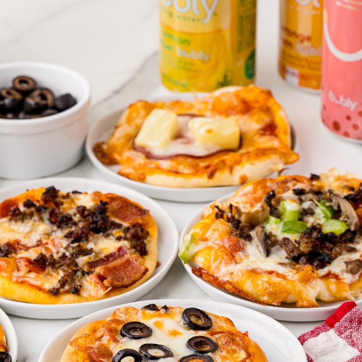 4 Rhodes Dinner Rolls mini pizzas on individual plates on a on a white marble countertop with 3 soda cans and a bowl of black olives in the back and a checkered red and white napkin in the front