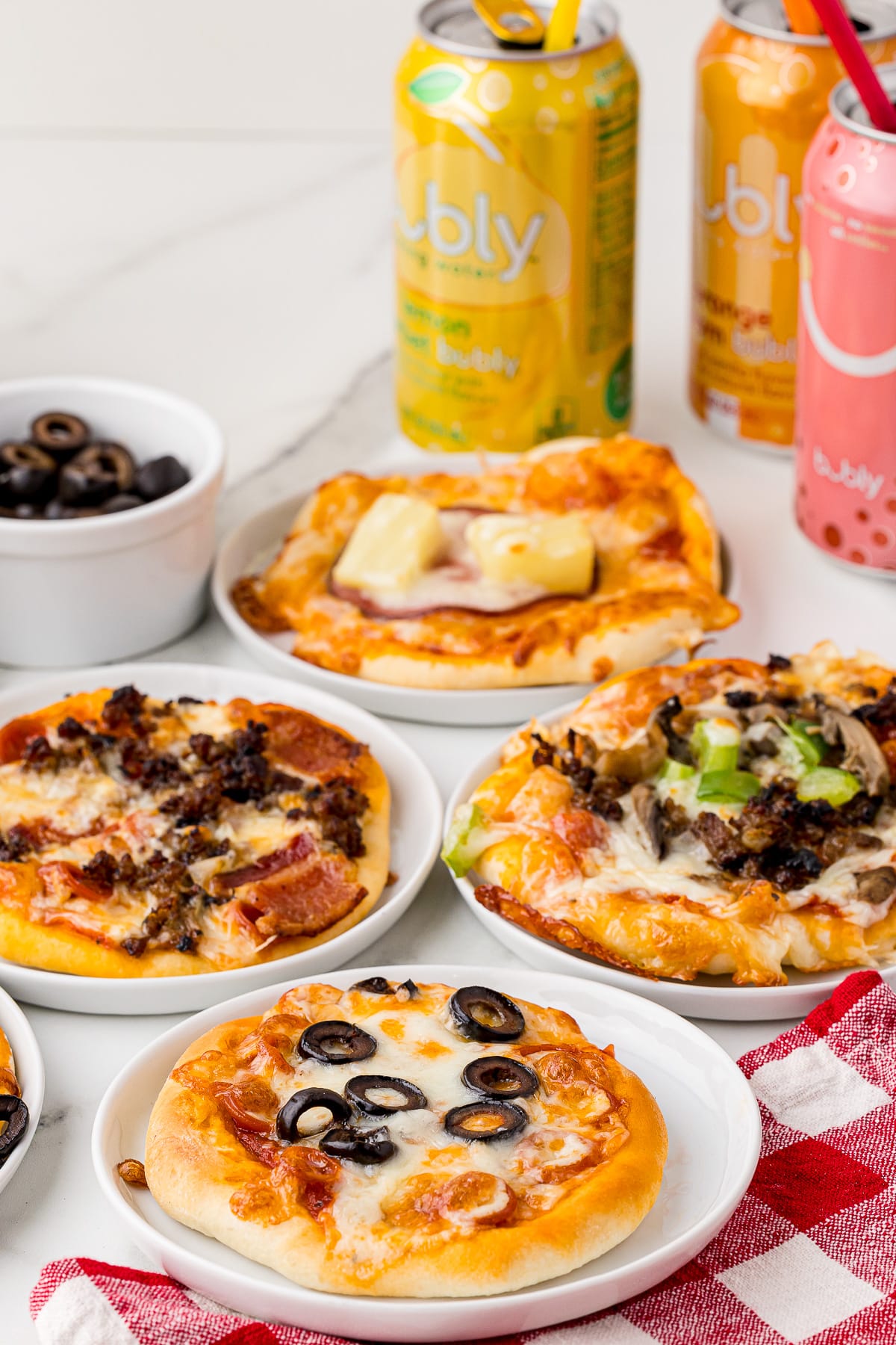 4 mini pizzas on individual plates on a on a white marble countertop with 3 soda cans and a bowl of black olives in the back and a checkered red and white napkin in the front