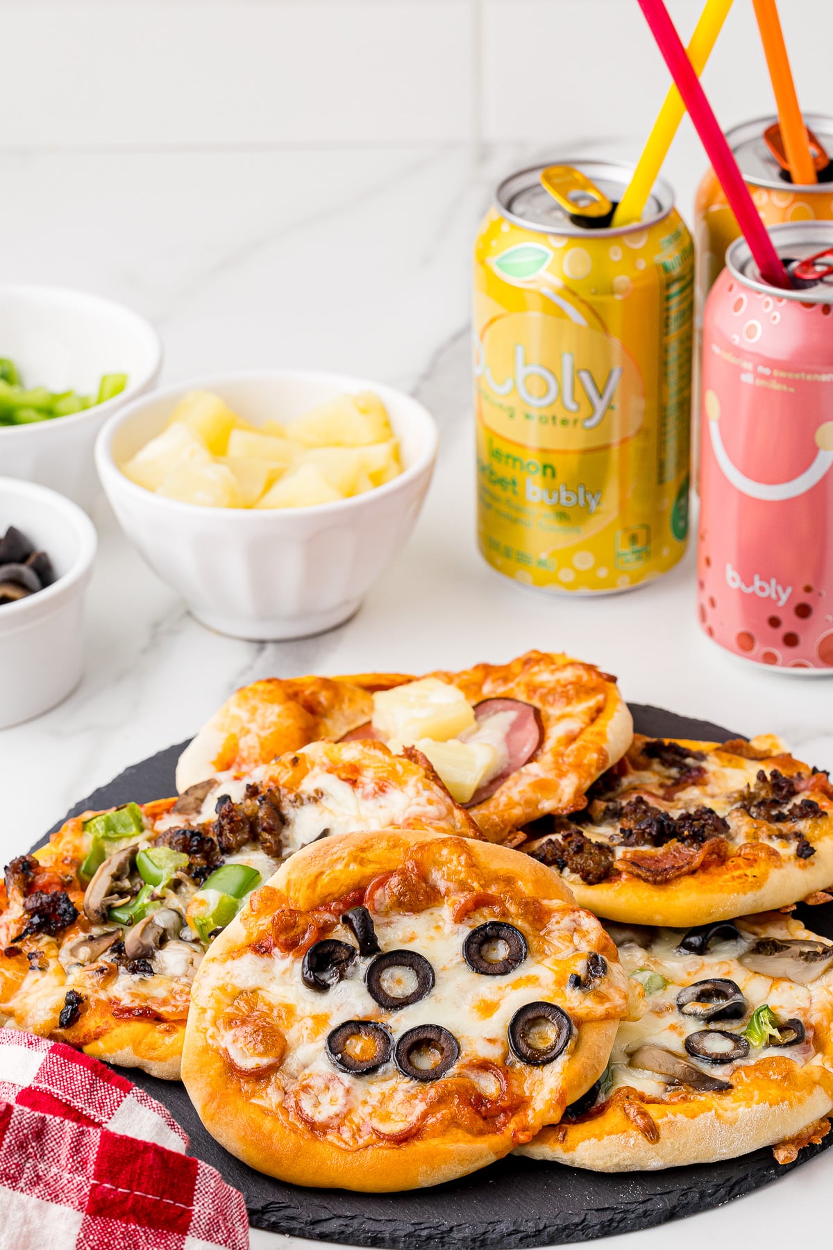 5 baked mini pizzas on a round slate plater with bubly soda cans and bowls full of toppings in the background, all on a white marble countertop