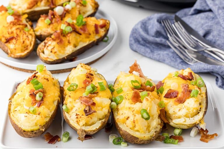 four twice baked potatoes on a white serving dish on a white counter with extra potatoes, a blue napkin and some forks and knives