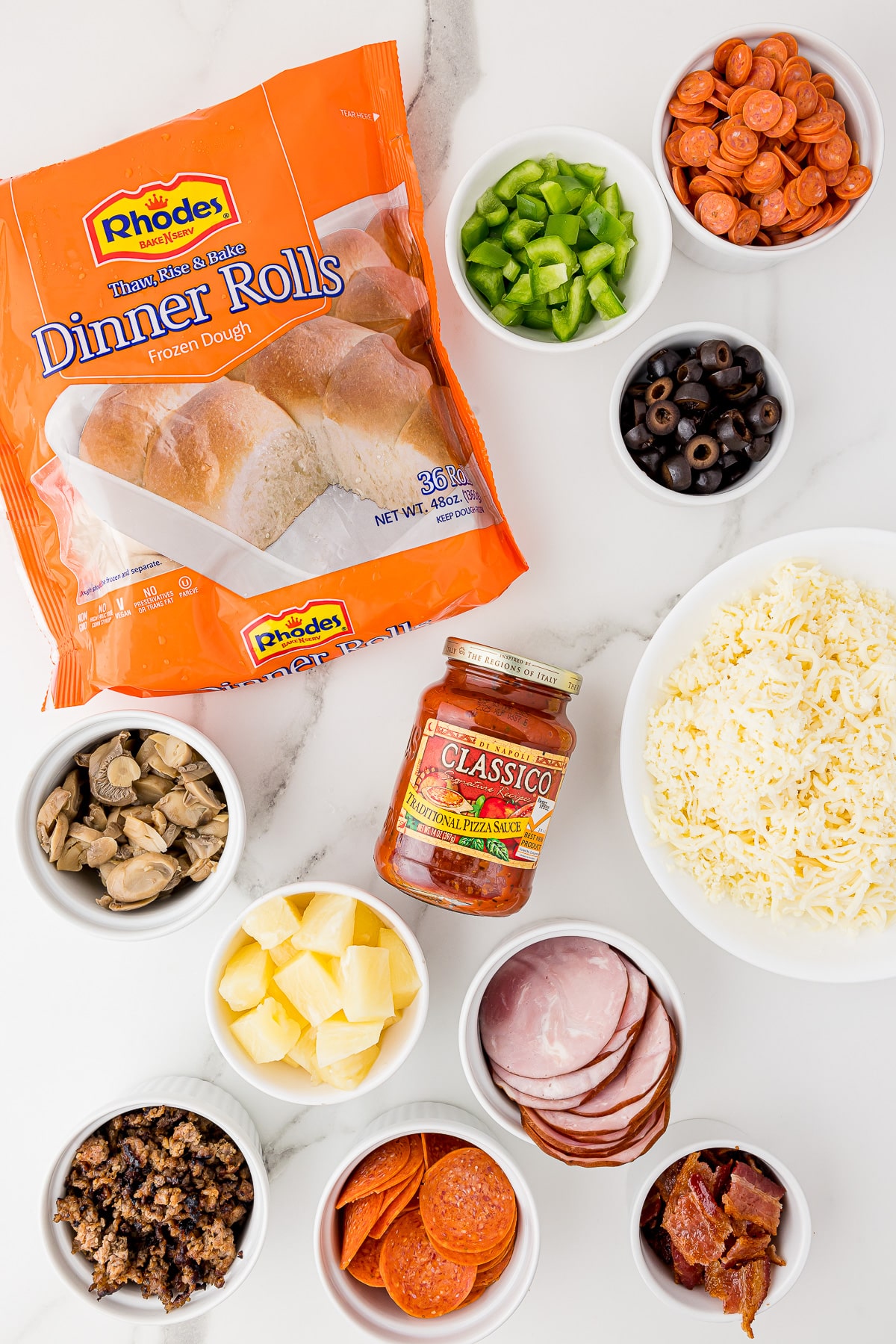ingredients for mini pizzas on a white marble countertop, including meats, olives, cheese, a package of Rhodes frozen dinner rolls and Classico traditional pizza sauce