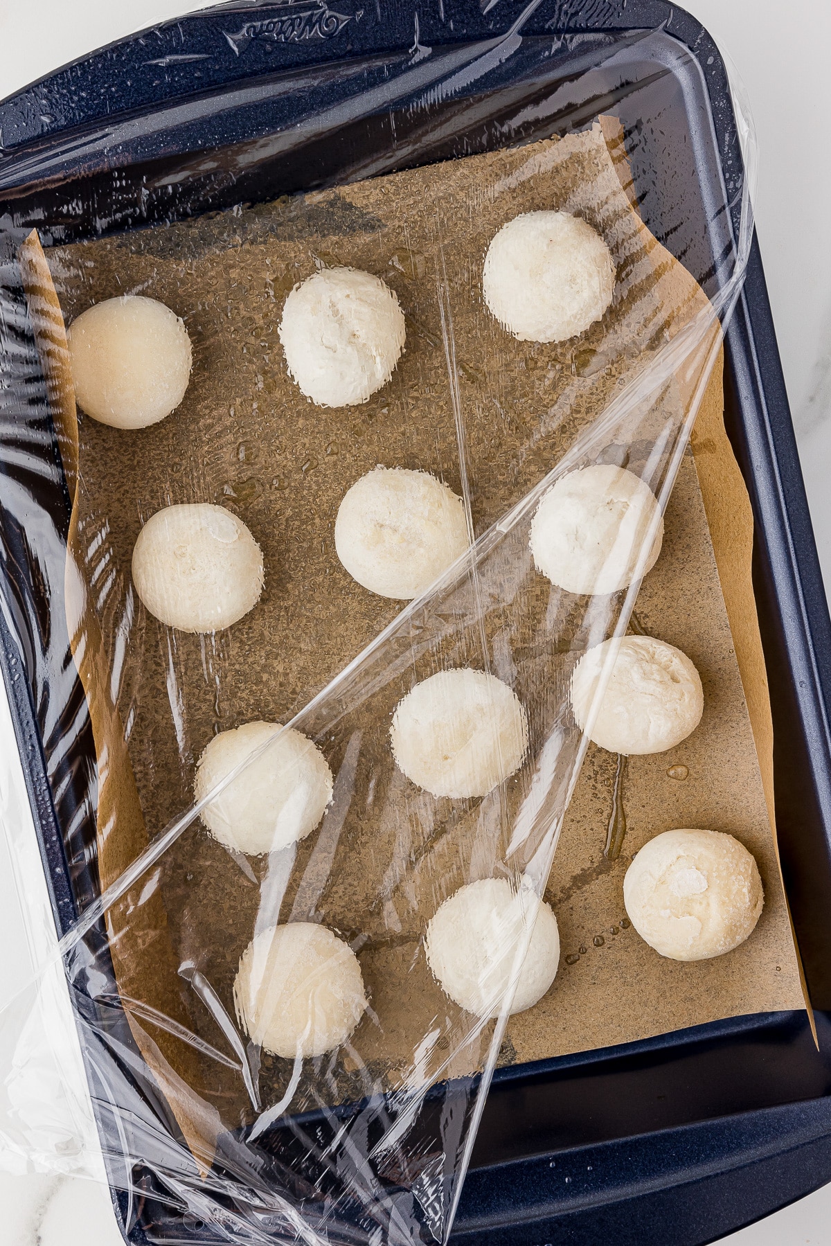 12 frozen Rhodes dinner rolls in a 9x13 Wilton cake pan, rolls are on parchment paper and are partially covered in plastic wrap