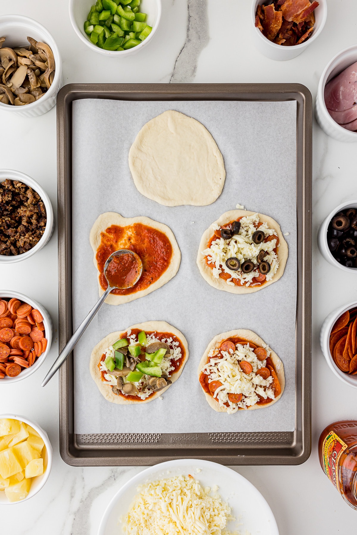cookie sheet covered in parchment paper with five mini pizzas being prepared, with topping surrounding the cookie sheet, all on a white marble countertop