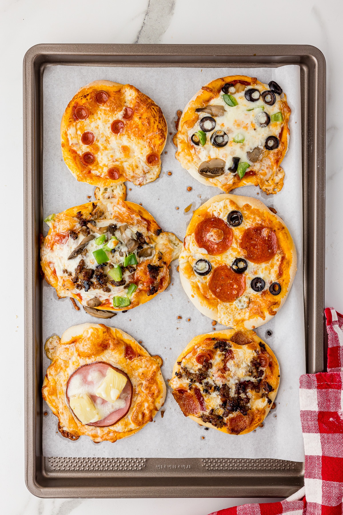 six mini pizzas on a cookie sheet with parchment paper and a checkered red and white napkin all on a white marble countertop