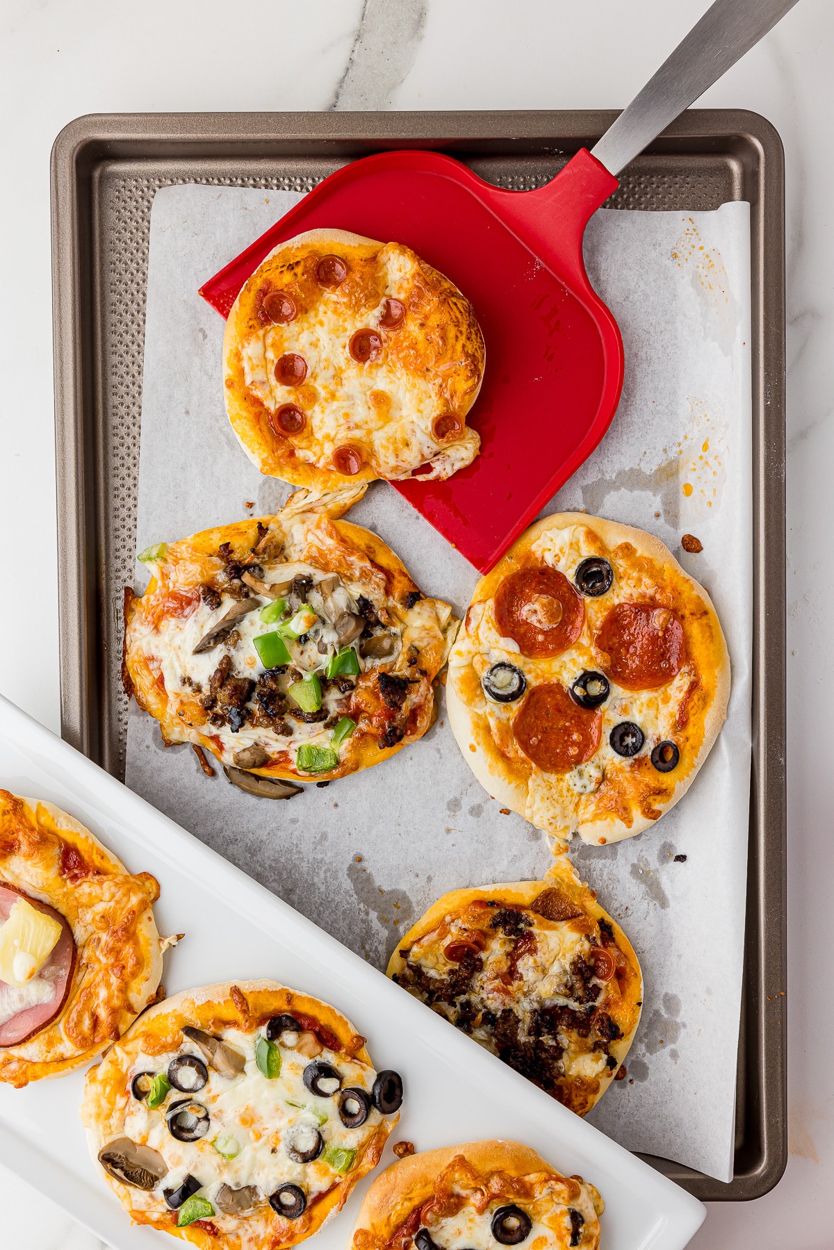 cookie sheet with 4 mini pizzas, one being lifted out of the pan with an oversized red Wilton spatula