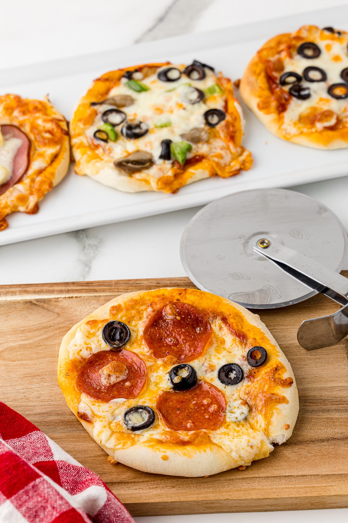 mini pizza on a cutting board with a red checkered napkin and a pizza cutter, with 3 mini pizzas in the background, all on a white marble countertop