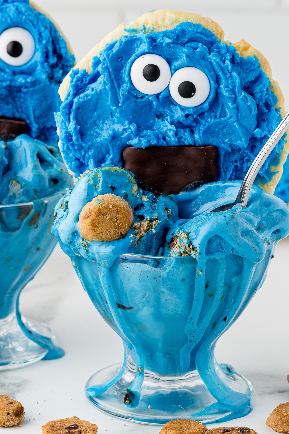 Blue cookie monster ice cream in a sundae dish with a cookie monster cookie and scattered mini chips ahoy on a white marble countertop