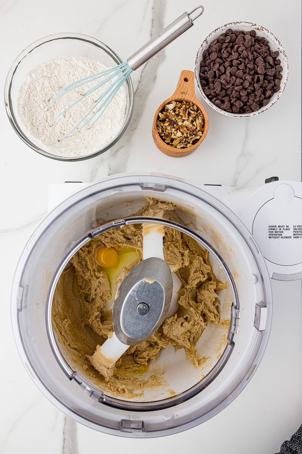 Bosch mixer with cookie dough on a white marble countertop with chocolate chips, walnuts and flour in bowls in the background