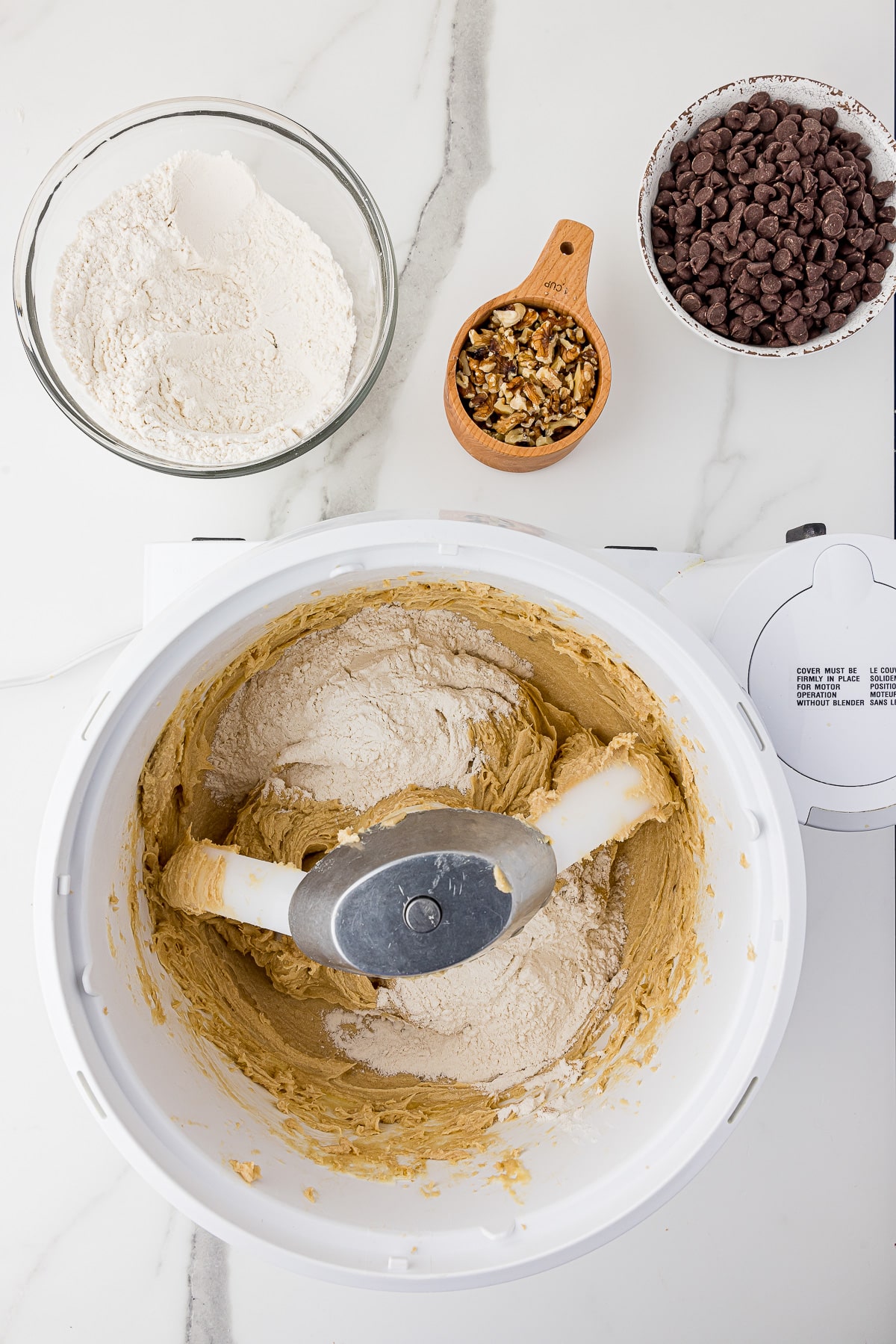 Chocolate chip cookie dough in a bosch mixer with a bowl of chocolate chips and a wooden measuring cup filled with walnuts and a glass bowl with flour in the back all on a white marble countertop