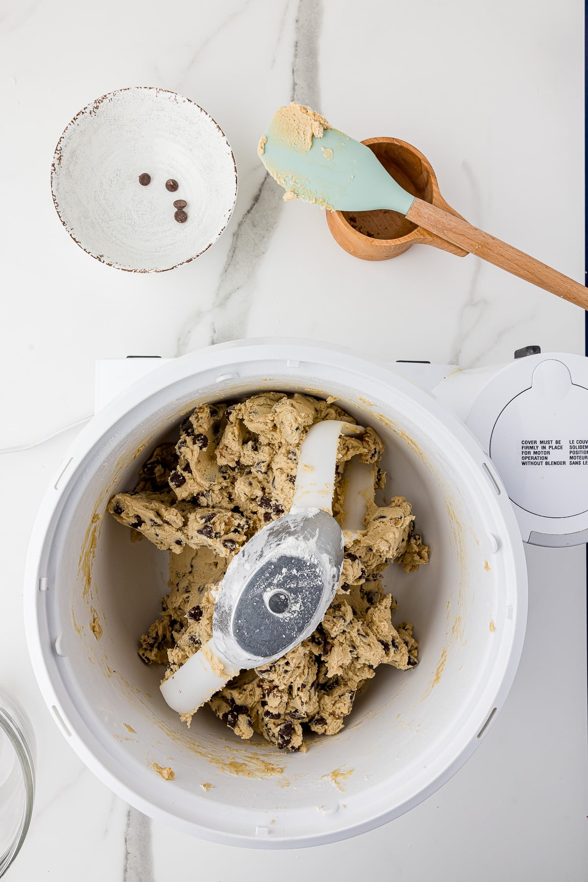 Chocolate chip cookie dough in a bosch mixer with a bowl of chocolate chips in the back and a teal spatula on top of a wooden measuring cup all on a white marble countertop