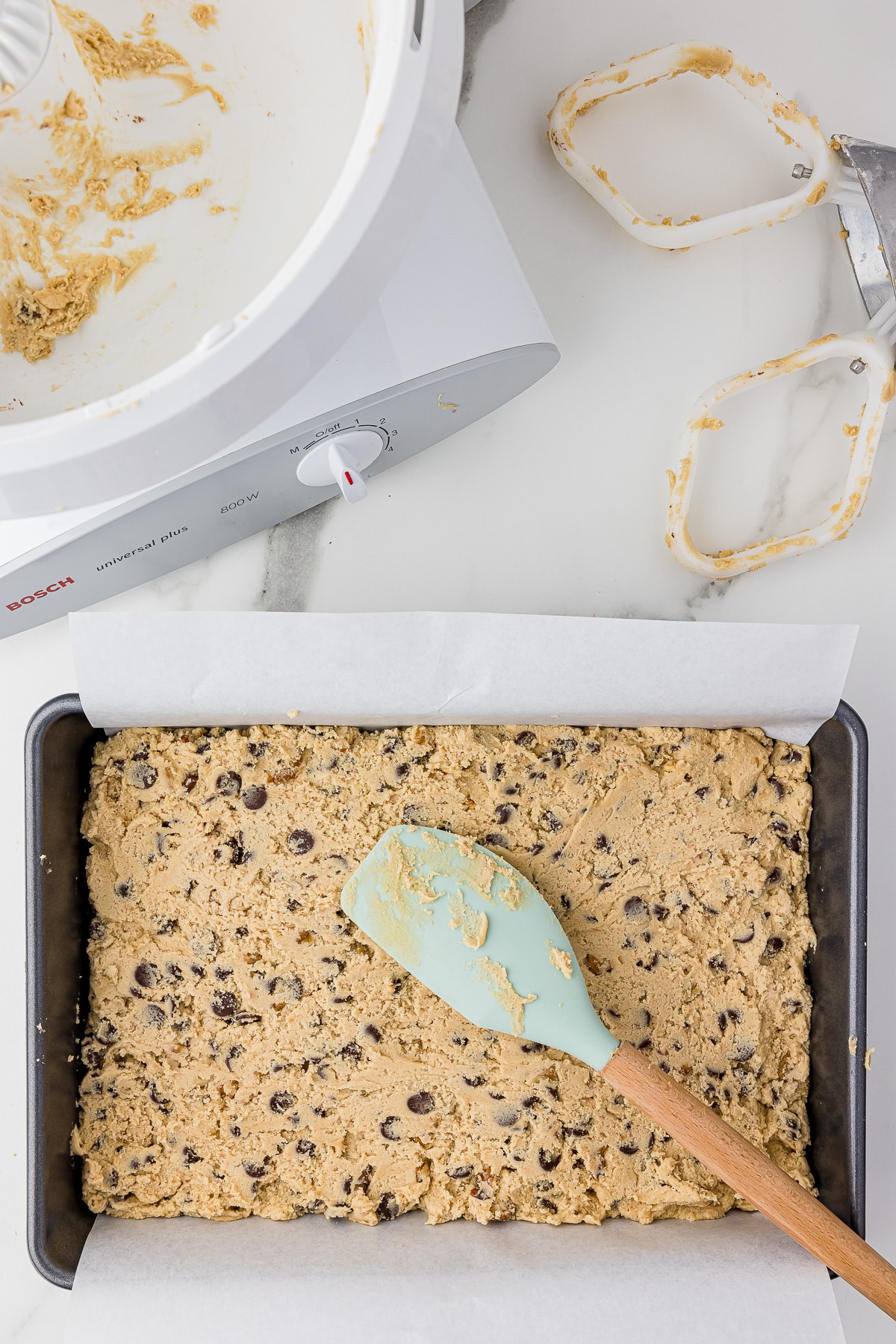 cookie dough in a 9x13 parchment lines pan with a teal spatula on top with a bosch mixer and its cookie paddles in the back, all on a white marble countertop