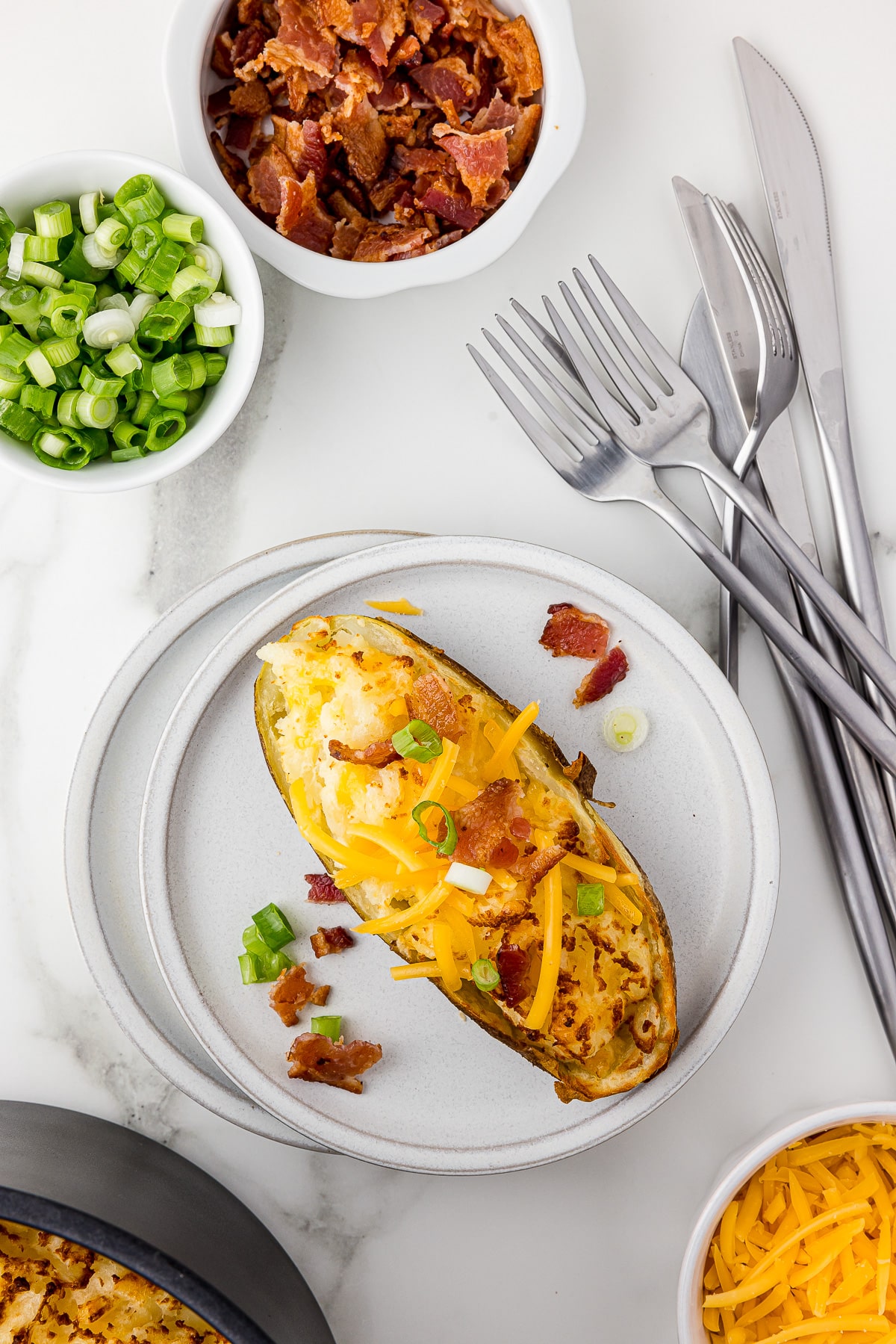 Twice baked potato on a white plate with forks and three knives on a white marble counter with a bowl of bacon bits, a bowl of sliced green onions, and a bowl of shredded cheddar cheese