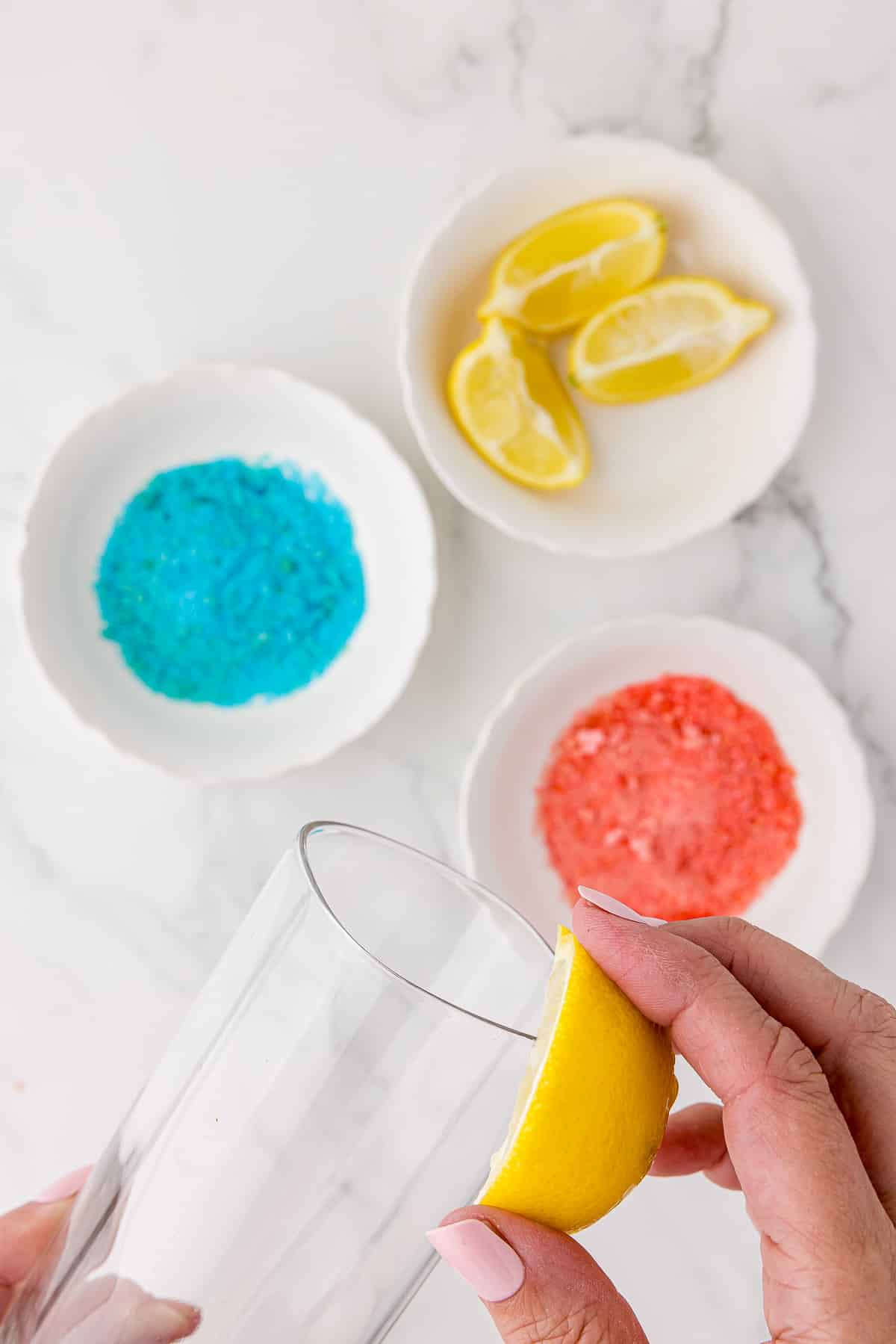Rubbing a lemon slice on the entire upper rim of a tall, square drinking glass. Also a small white bowl with blue powdery candy, a small white bowl with red powdery like candy, and a small white bowl with sliced lemon