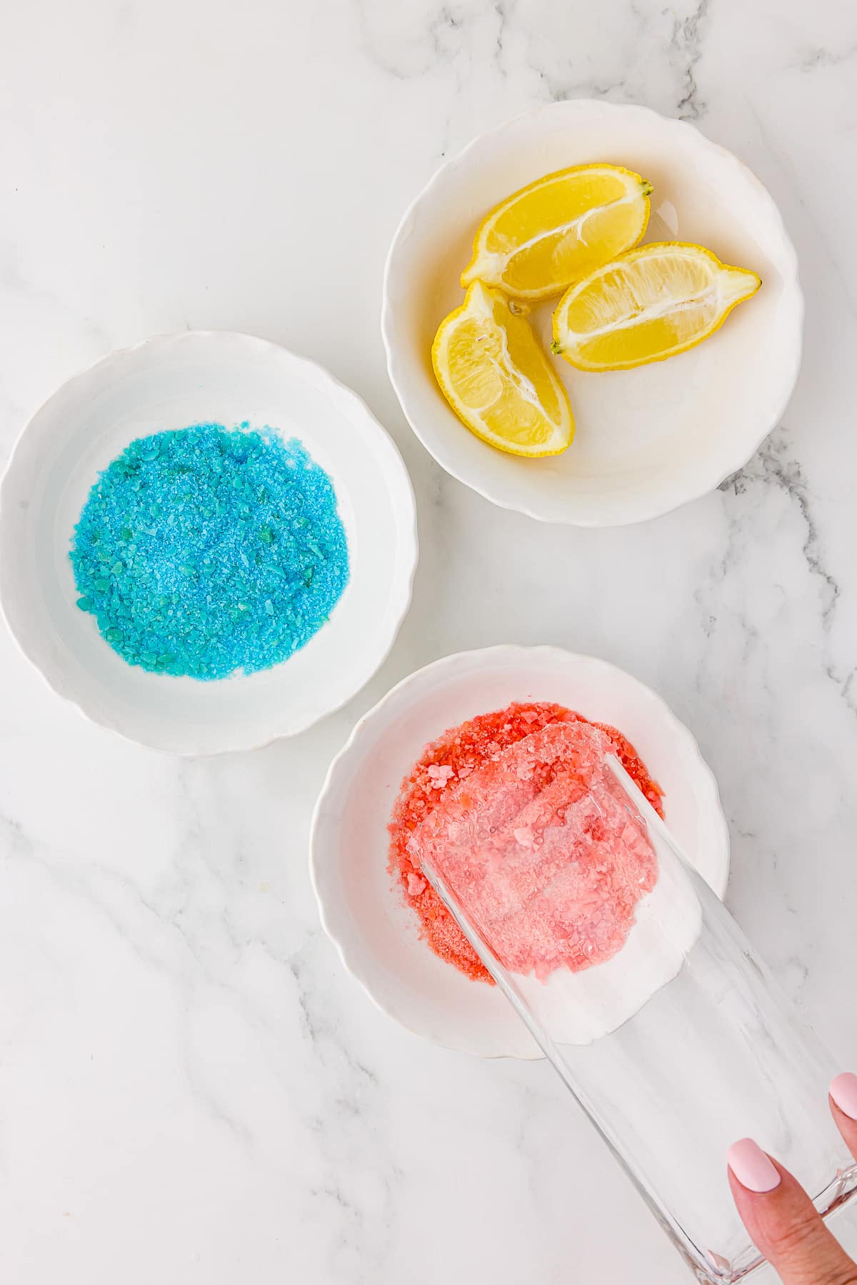 Dipping a tall square glass, the upper rim of which has been covered with lemon juice, into a small white bowl of red pop rock like candy. Also, a small, whtie bowl of blue pop rock candy and a small, white bowl of sliced lemon