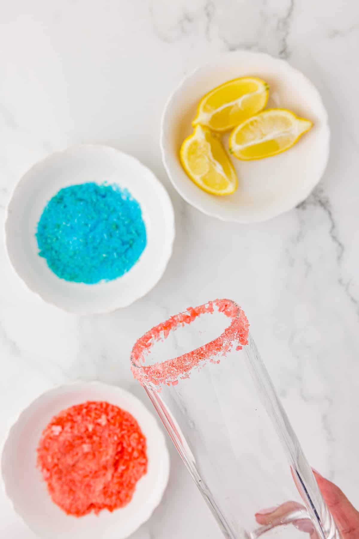 Two small white bowls of pop rock like candy, one with red candy and one with blue candy, and a small white bowl of sliced lemons, and a square glass, the upper rim of which was dipped in lemon juice and then into the red candy