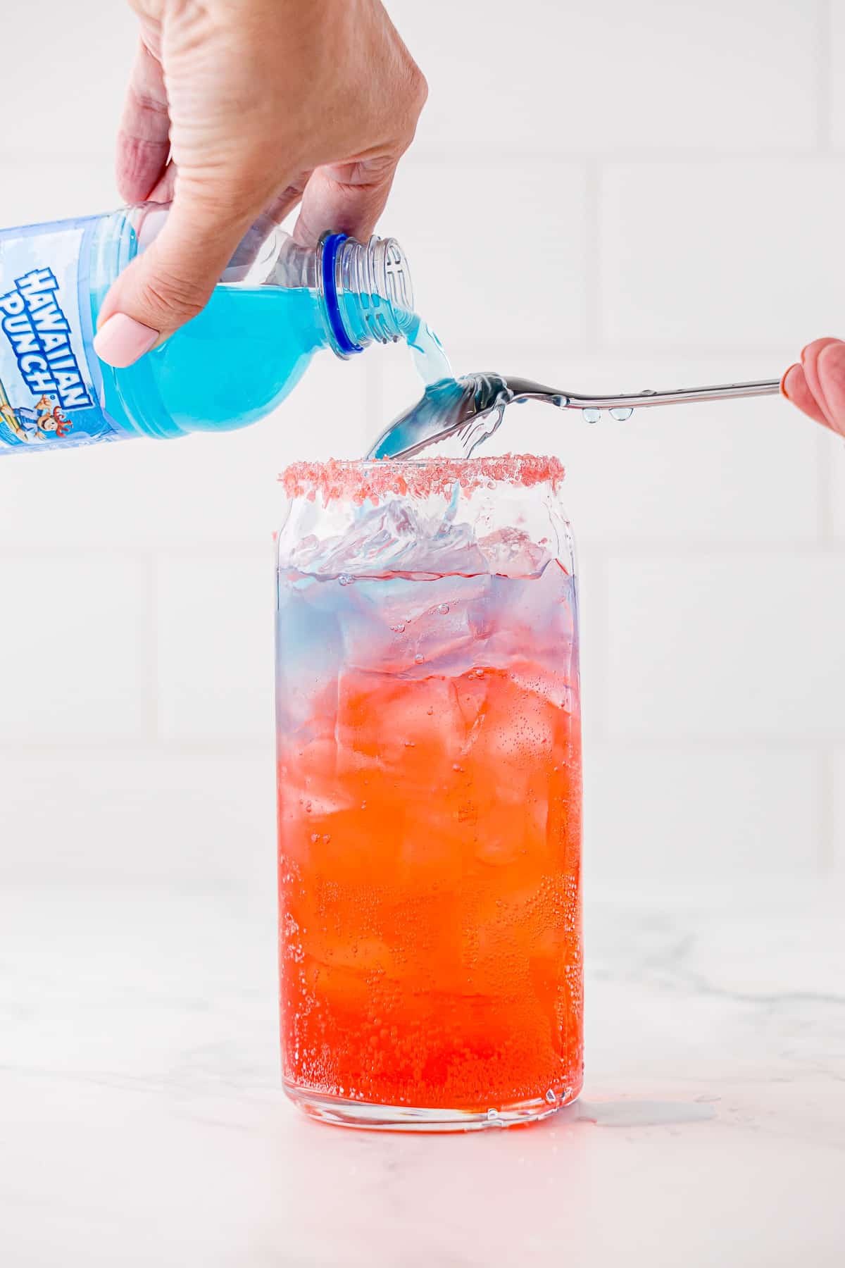 Adding the top blue Hawaiian Punch layer to a tall, round drinking glass that already has red and white layers of liquid in it.