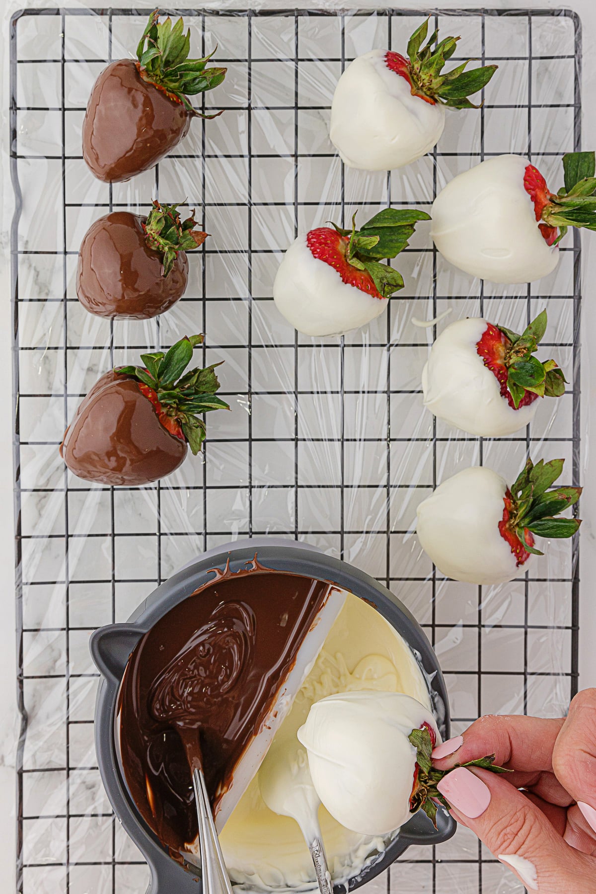 3 strawberries dipped in dark chocolate and 5 strawberries dipped in white chocolate on a rectangular metal rack, and 1 more strawberry in hand recently dipped in white chocolate