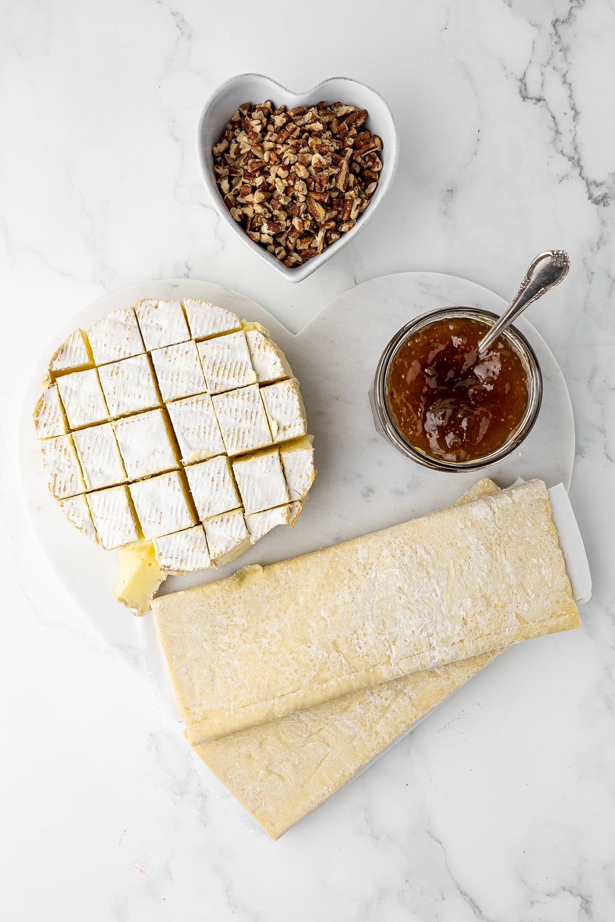white marble countertop with a heat shaped cutting board in the middle. A cubed wheel of brie, a jar of fig jam, and two sheets of folded puff pastry are on the cutting board and a heart shaped dish hold chopped walnuts.
